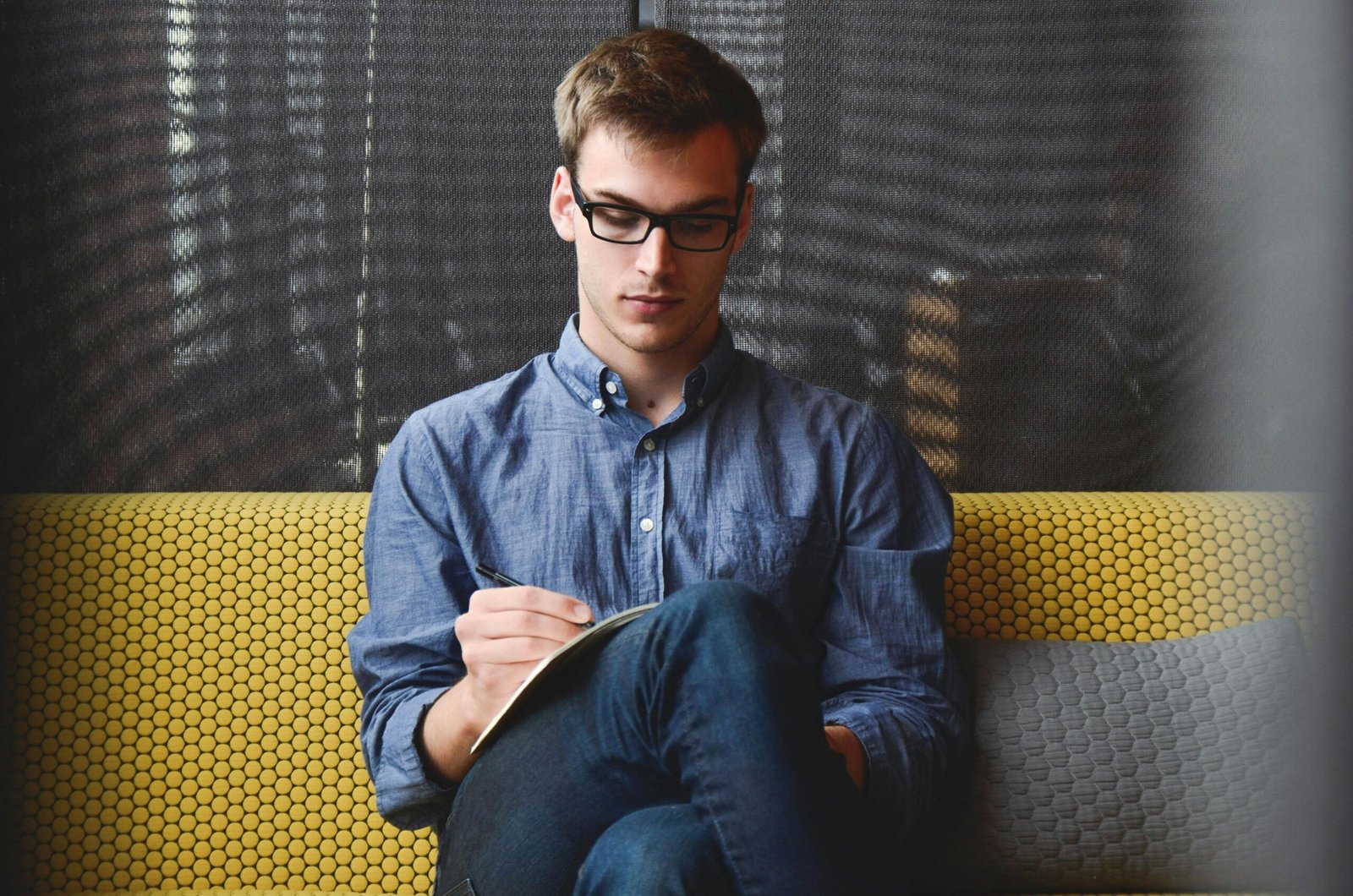 Web Martín Cuesta A young man in glasses writes in a notebook while sitting on a stylish couch indoors.